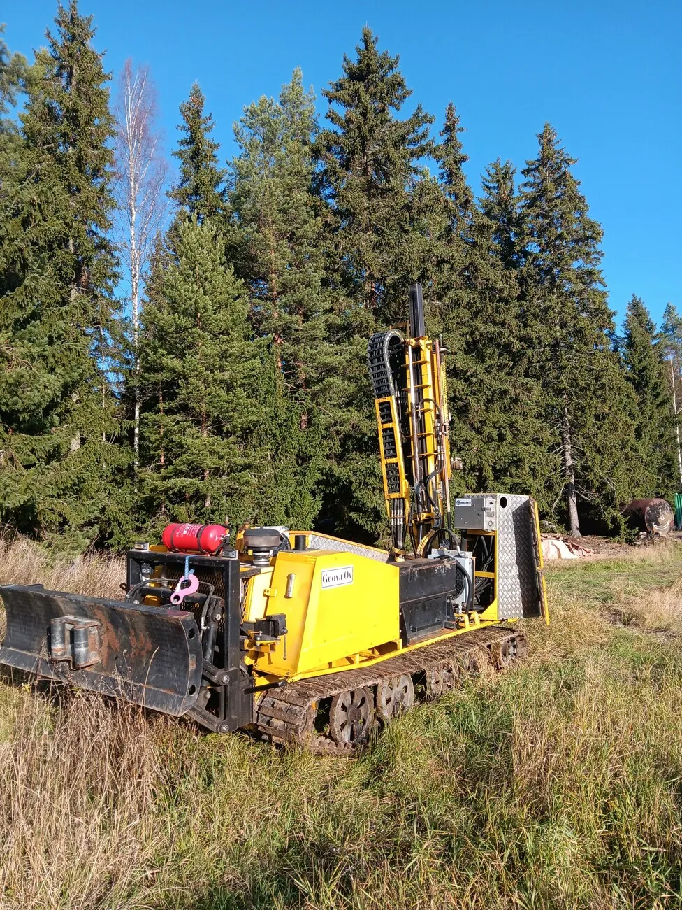 Keltainen tela-alustainen porauslaite seisoo ruohoisella pellolla metsän reunassa maaperätutkimuksia varten.