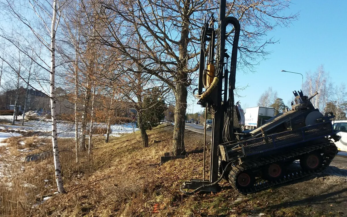 Porausvaunu suorittaa maaperätutkimusta ruohoisella rinteellä tien vieressä, taustalla puita ja rakennuksia.