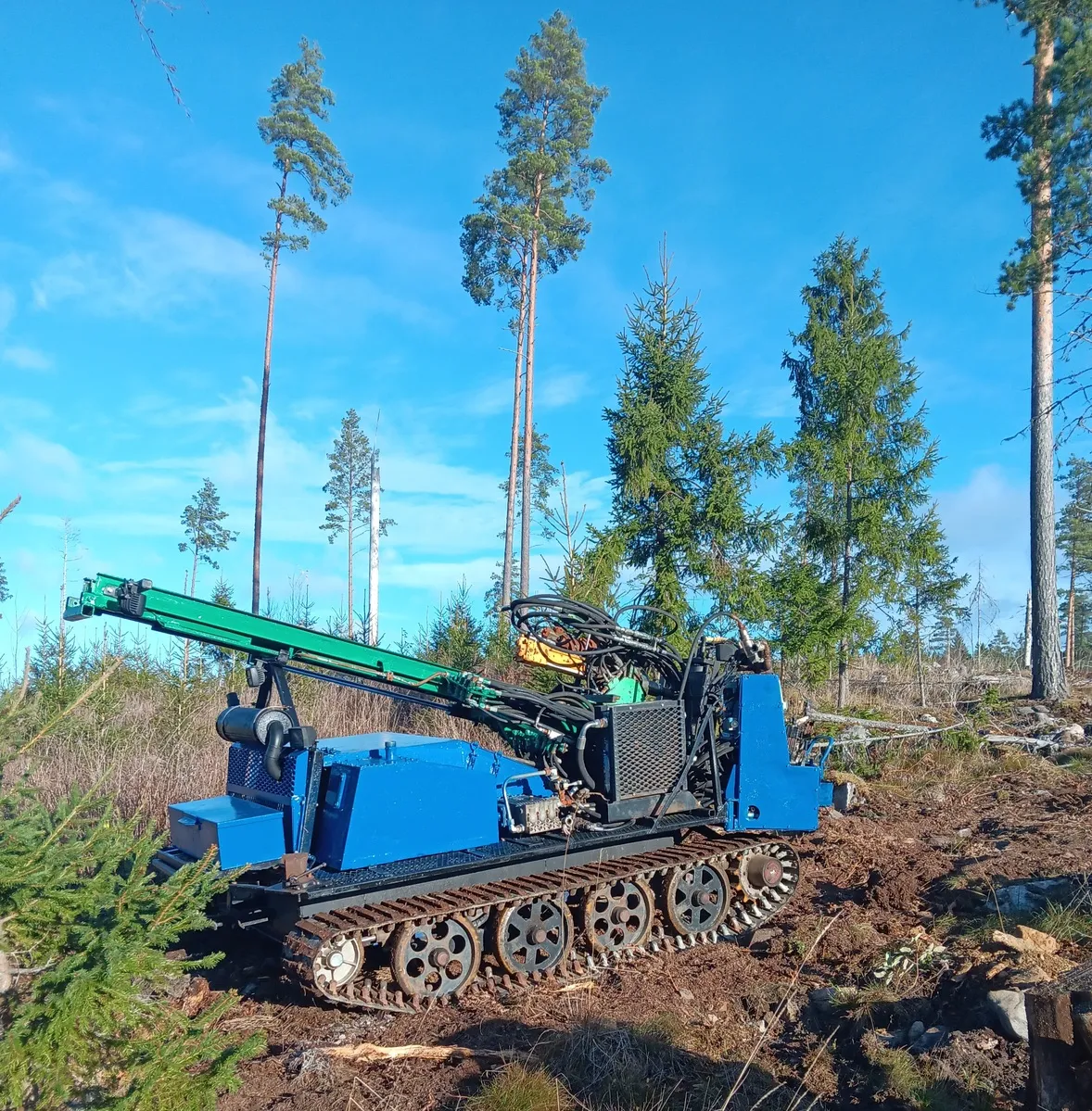 Sininen telaketjuporavaunu tekee maaperätutkimusta metsäisessä maastossa, jossa on korkeita mäntyjä ja kuusia.